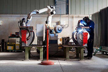 Man and robotic machine work together inside industrial building. The mechanical arm performs welds on metal components assisted by a worker who in turn manages welds manually.